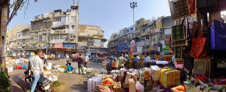 Busy street market in Delhi showing vendors with colorful goods, sacks of merchandise, hanging textiles and bags, multi-story buildings with shop signs, people shopping and walking, motorcycles parked along the street