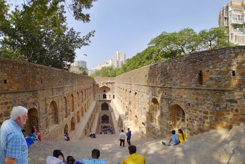 Historical stone stepwell in Delhi with multiple levels of arched niches along sandstone walls, visitors sitting and walking on descending steps, modern buildings visible in background against blue sky