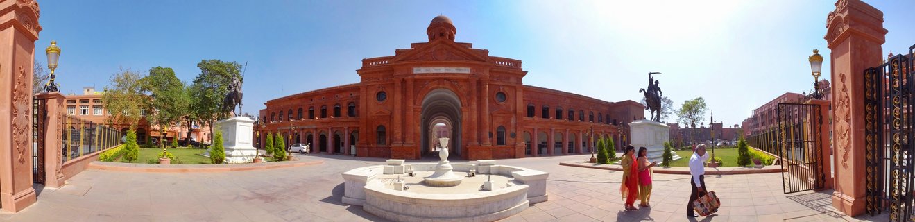 Red sandstone colonial building with central arched entrance and dome, flanked by two equestrian statues on white pedestals, white marble fountain in foreground, people in colorful saris walking on paved courtyard, Amritsar India