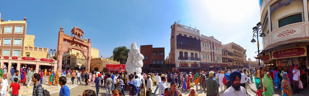 Wide panoramic street view showing a large crowd of people in colorful clothing walking through a plaza in Amritsar, India, with a white sculpture in the center and multi-story buildings with shops surrounding the area under a bright blue sky