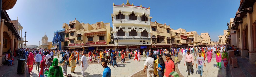 A wide street scene in Amritsar, India showing a crowded plaza with people in colorful traditional Indian clothing walking between ornate cream and yellow buildings with decorative balconies under a clear blue sky