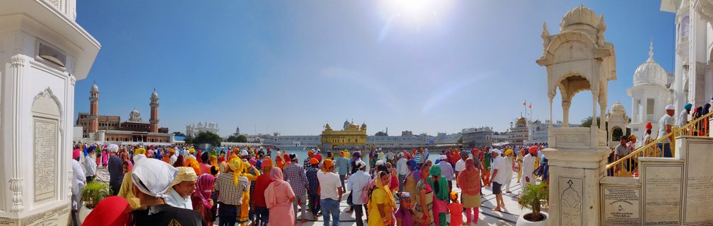 Crowds of Sikh pilgrims in colorful turbans walking around the Golden Temple complex in Amritsar with the gold-plated sanctum visible across the sacred pool under bright blue sky