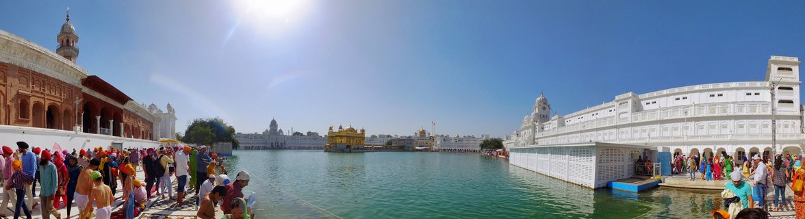 Wide panoramic photograph showing the Golden Temple in the center of a holy water tank with crowds of people walking along marble pathways on both sides under bright sunlight