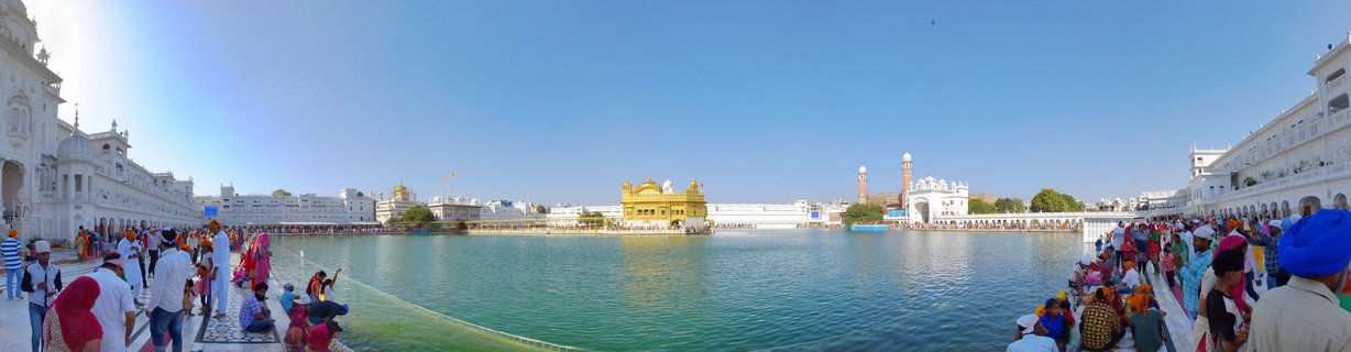 Panoramic view of the Golden Temple in Amritsar showing the gold-plated central shrine surrounded by a large sacred water tank with hundreds of visitors along the marble walkways under a clear blue sky