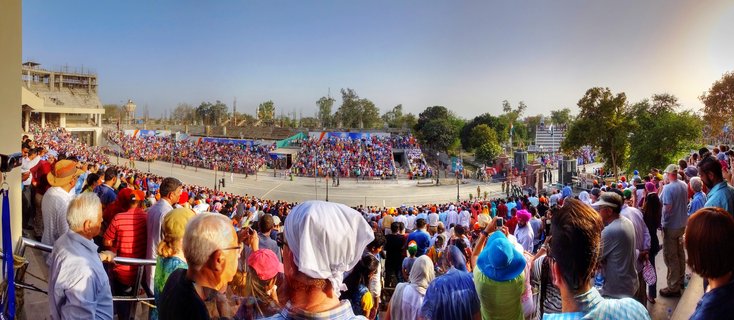 Panoramic view of large crowd seated in stadium bleachers watching the Wagah Border ceremony with parade ground in center and clear blue sky above