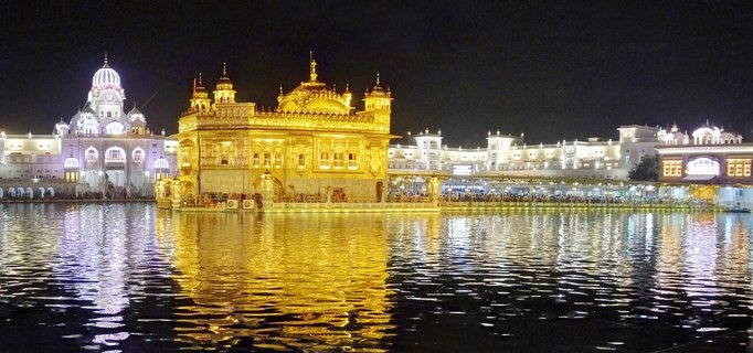 Night photograph of the Golden Temple in Amritsar, India, showing the illuminated golden building reflected in the surrounding sacred water pool with white marble buildings in the background