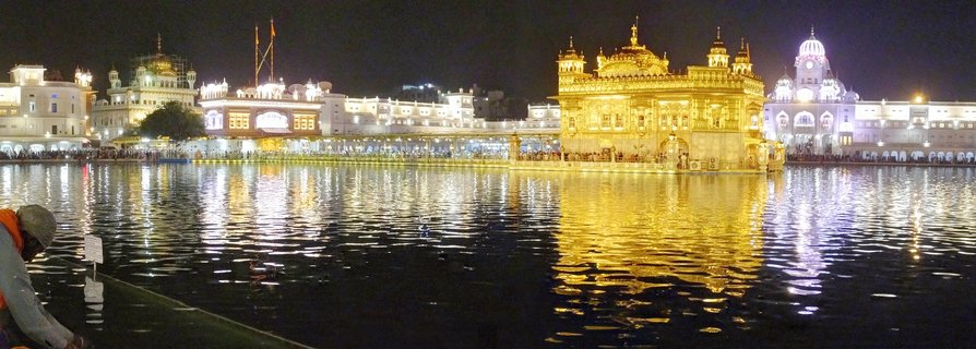 Night photograph of the Golden Temple in Amritsar, India, showing the gold-plated Sikh shrine illuminated and reflected in the surrounding sacred water pool with white buildings and visitors visible around the complex