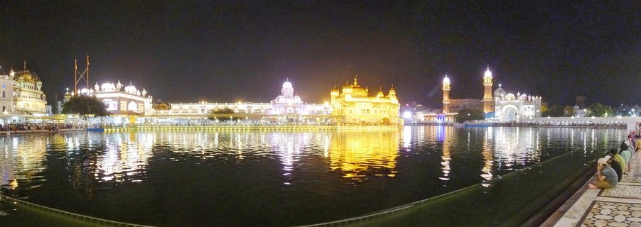 Nighttime panoramic view of the Golden Temple in Amritsar, India, with the illuminated golden building reflected in the dark water of the sacred pool, pilgrims visible sitting along the marble walkway on the right side