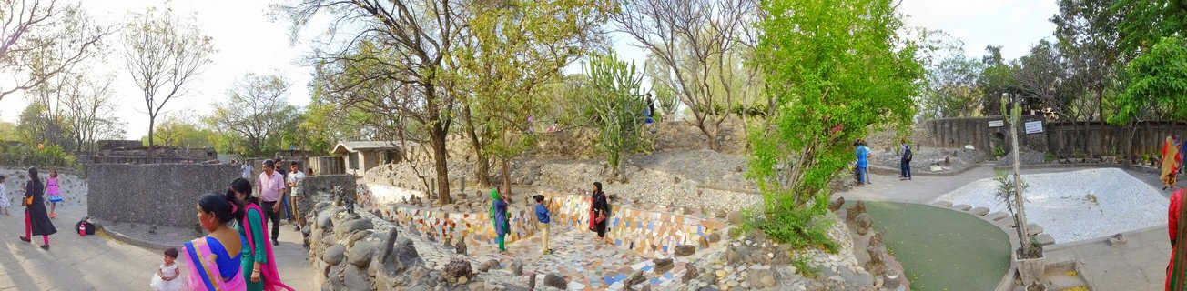 Panoramic view of visitors walking through Nek Chand's Rock Garden in Chandigarh, India, featuring colorful mosaic tile walls, terraced pathways, trees, and a small green pond