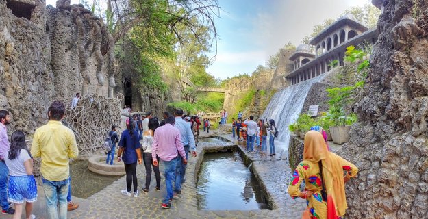 Wide outdoor scene showing tourists walking on stone pathways beside an artificial waterfall at Rock Garden in Chandigarh, India, with unique rock sculptures and domed buildings surrounded by green trees