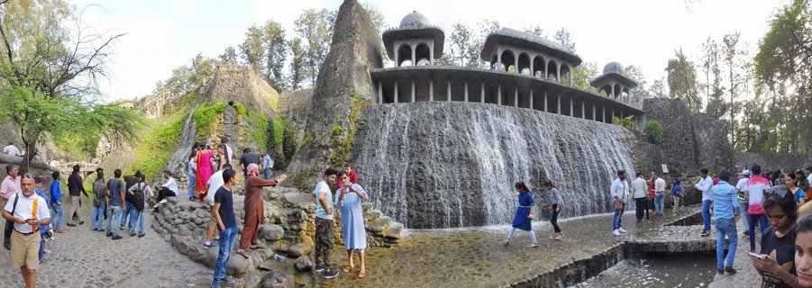 Wide panoramic view of tourists walking around an artificial waterfall with domed stone pavilions at Rock Garden in Chandigarh, India