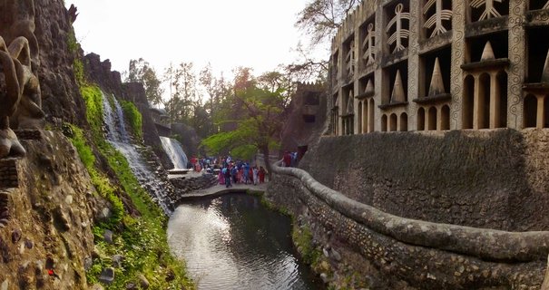 Outdoor scene showing waterfalls flowing down rocky terrain on the left, a pond in the center foreground, and multi-level concrete architecture with geometric patterns on the right, with visitors walking on pathways between the features