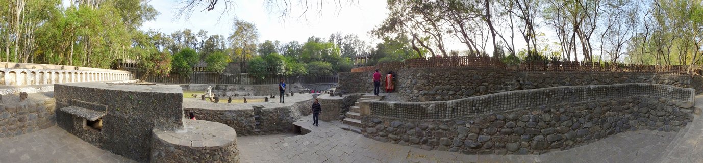 Panoramic view of ancient stone ruins with multiple terraced levels, stone walls, and visitors walking on pathways surrounded by trees