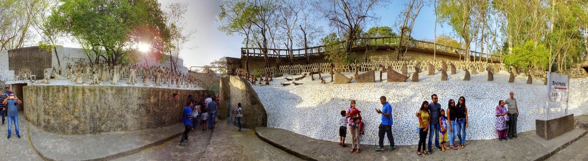 Panoramic view of the Rock Garden in Chandigarh showing white mosaic tiled walls, recycled material sculptures, visitors walking on stone pathways, and trees surrounding the outdoor art installation
