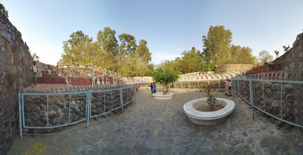 Outdoor sculpture garden with stone walls displaying various statues and sculptures, metal fencing, circular white brick planters with small trees, and visitors walking on paved courtyard under clear blue sky
