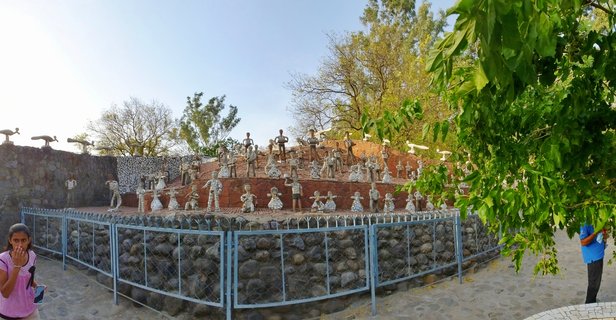 Panoramic view of an archaeological site in Chandigarh showing ancient stone stupa ruins with sculptural elements, surrounded by metal railings and trees with visitors walking on sandy pathways