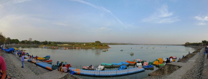 Wide panoramic view of Sukhna Lake in Chandigarh showing colorful paddle boats docked at blue floating platforms, visitors walking along stone embankments, calm water with boats scattered across the surface, and a small tree-covered island in the distance under a blue sky with wispy clouds