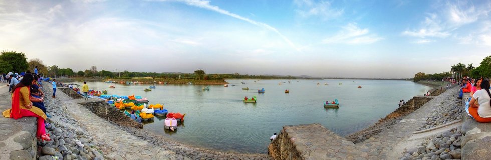 Panoramic view of Sukhna Lake in Chandigarh showing colorful paddle boats on water with people sitting on stone embankments on both sides
