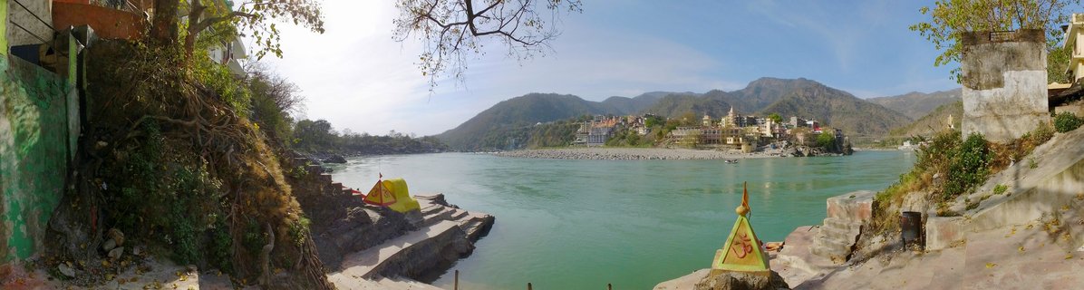 Wide panoramic view of the Ganges River in Rishikesh showing stone steps leading to green water, buildings and ashrams on both banks, and mountains in the distance under a bright sky