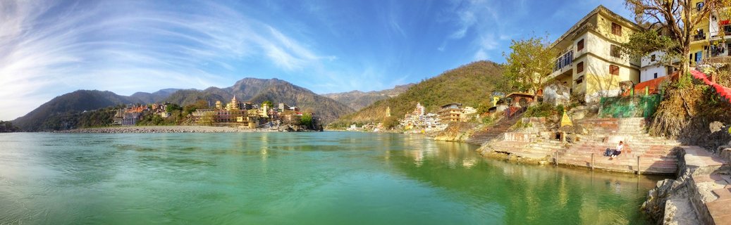 Panoramic view of the Ganges River in Rishikesh showing turquoise water, stone steps leading to the river, colorful temple buildings along the banks, and mountains in the background under a blue sky