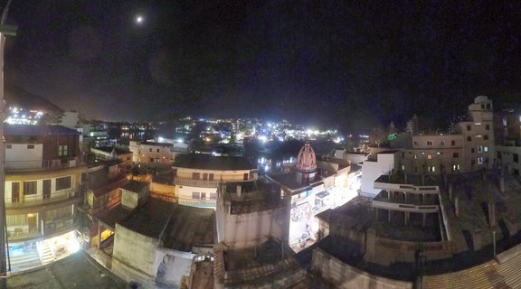 Night panoramic photograph of Rishikesh city showing multiple buildings with flat roofs, illuminated windows, a temple dome, and moon in dark sky
