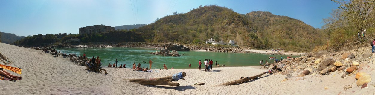People bathing and relaxing on a sandy beach along the turquoise Ganges River in Rishikesh, India, with forested hills and buildings in the background under clear blue sky