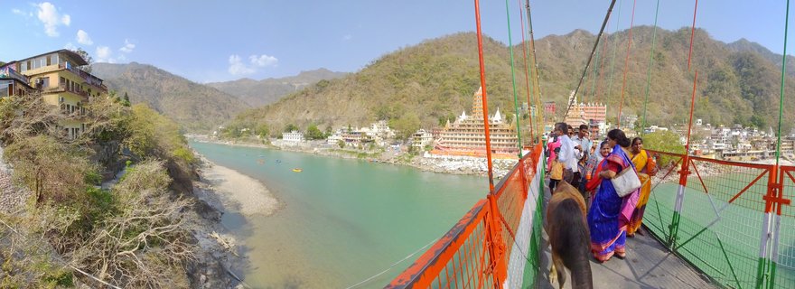 People walking on an orange suspension bridge over turquoise river with temples, buildings, and mountains in Rishikesh, India
