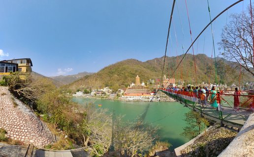 Wide view of Ram Jhula suspension bridge with colorful cables spanning the Ganges River in Rishikesh, India. People in traditional clothing walk across the bridge. Hindu temples with orange and cream architecture line the far riverbank. Forested hills rise in the background under a clear blue sky.