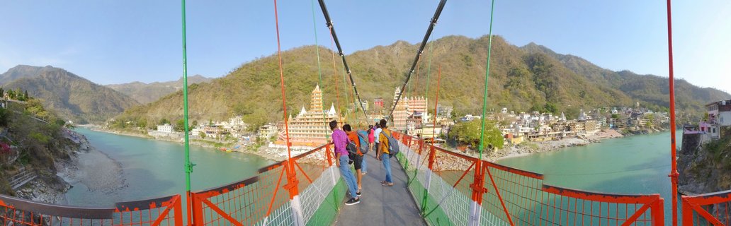 People walking on a colorful suspension bridge with orange and green cables over a wide turquoise river, with Hindu temples and mountains in the background in Rishikesh, India