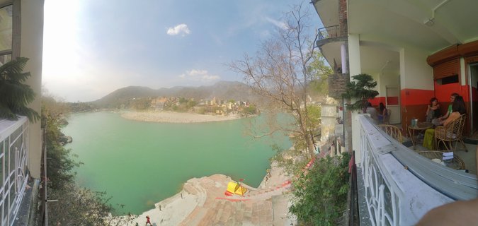 Wide panoramic photograph showing the Ganges River in Rishikesh, India, viewed from a cafe balcony with people seated at tables, stone steps leading to turquoise water, buildings and temples on far bank, and mountains in the background under blue sky