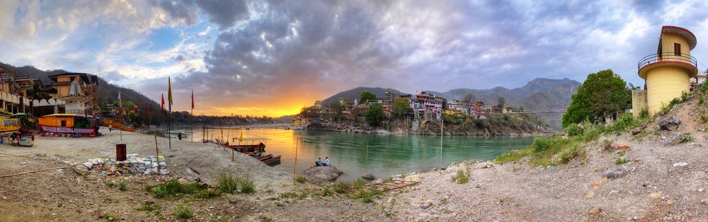 Panoramic view of the Ganges River at sunset in Rishikesh, India, showing riverside buildings, boats on the shore, a yellow tower structure, and mountains under cloudy skies with golden sunset light