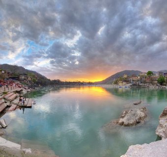 Wide panoramic view of the Ganges River in Rishikesh, India at sunset with stone ghats, pilgrims, temples, and Himalayan foothills under dramatic cloudy sky