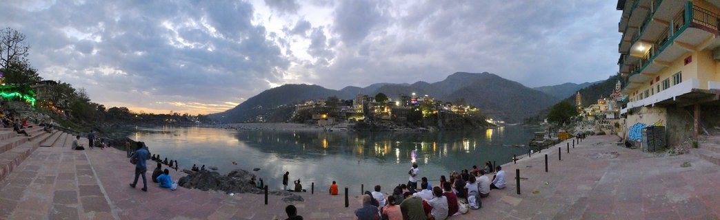 Wide view of people sitting and standing on stone steps along the Ganges River in Rishikesh at dusk, with temple buildings, mountains in the background, and cloudy twilight sky