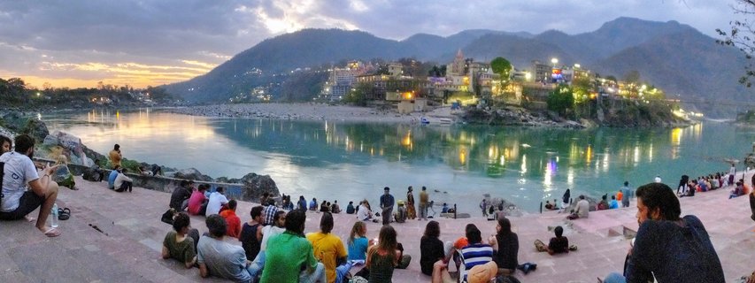 Wide panoramic view of people sitting on stone riverfront steps at dusk, with the Ganges River flowing past illuminated buildings and temples built into a hillside, mountains visible in the background under a twilight sky