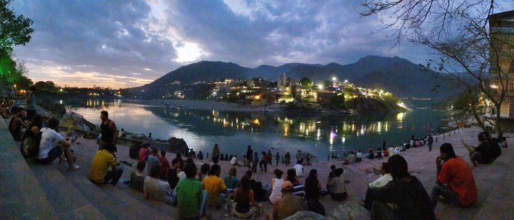 Panoramic view of people sitting on riverside steps along the Ganges River at dusk, with illuminated buildings on a hillside and mountains in the background under a cloudy twilight sky