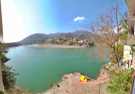 Aerial view of the Ganges River in Rishikesh showing turquoise water, stone ghats with people bathing, yellow tent, buildings on the far bank, and mountains in the background under blue sky.