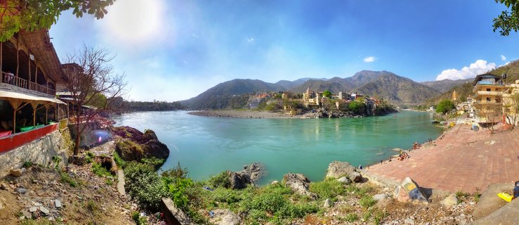 Panoramic view of the Ganges River in Rishikesh, India, with turquoise water, traditional buildings, stone steps, and mountains in the background under blue sky