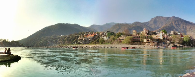 Wide river view showing red rafting boats on greenish-blue water with temple buildings and mountains in the background at Rishikesh, India