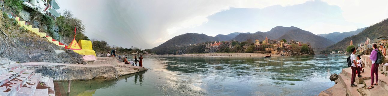 Wide panoramic view of the Ganges River in Rishikesh showing stone steps leading to water, people sitting on ghats, yellow religious structure with Om symbol, temple buildings across the river, and mountains in the background under cloudy sky