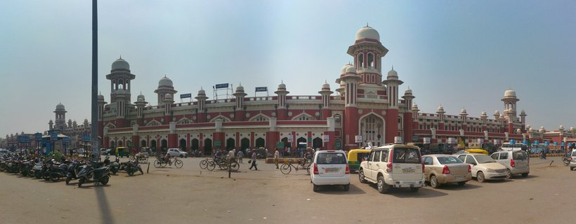 Wide view of Lucknow Charbagh Railway Station showing red brick building with multiple white domes and towers, vehicles and motorcycles parked in foreground, people walking, clear blue sky