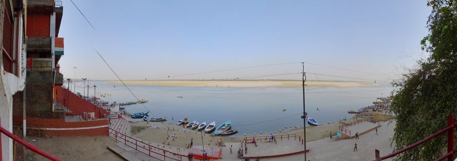 Wide view of Ganges River ghats in Varanasi with boats docked along the shore, stone steps leading to water, red buildings on the left, and people walking along the riverbank under a clear sky