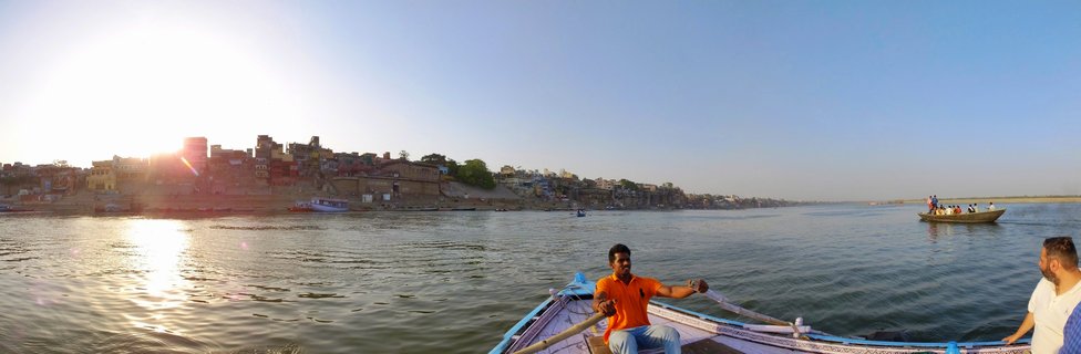 A boatman wearing an orange shirt rows a traditional wooden boat on the Ganges River in Varanasi, India, with historic ghats and buildings lining the riverbank under a clear sky during late afternoon sunlight.
