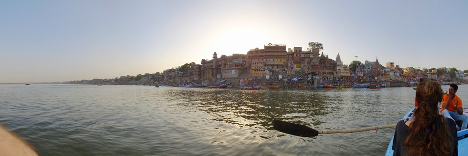 Wide panoramic photograph of Varanasi ghats along the Ganges River at sunset, with boats on the water and people visible in a boat in the foreground