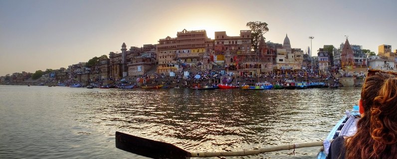 Panoramic view of Varanasi ghats along the Ganges River at sunset with ancient buildings, colorful boats, pilgrims on steps, and water reflections