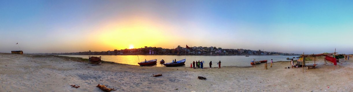 Panoramic sunset view of the Ganges River in Varanasi with wooden boats on sandy shore and people gathered near the water