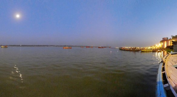 Panoramic view of the Ganges River in Varanasi at twilight with multiple wooden boats, illuminated ghats on the right bank, and the moon visible in the blue evening sky