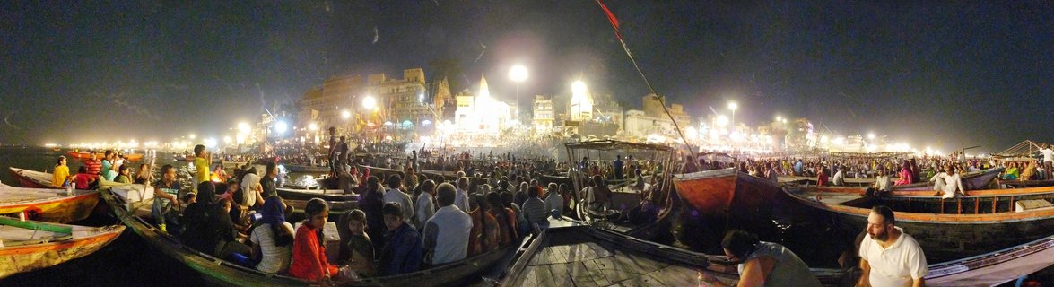 Night panoramic view of crowded wooden boats on the Ganges River with illuminated temple ghats and crowds during Ganga Aarti ceremony in Varanasi, India