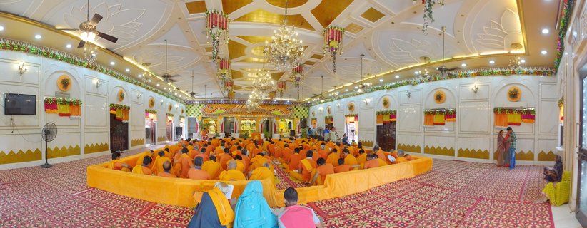 Interior view of a Hindu temple hall in Varanasi with hundreds of people in orange robes seated on patterned carpet, ornate white and gold ceiling with crystal chandeliers, decorative flower garlands along walls
