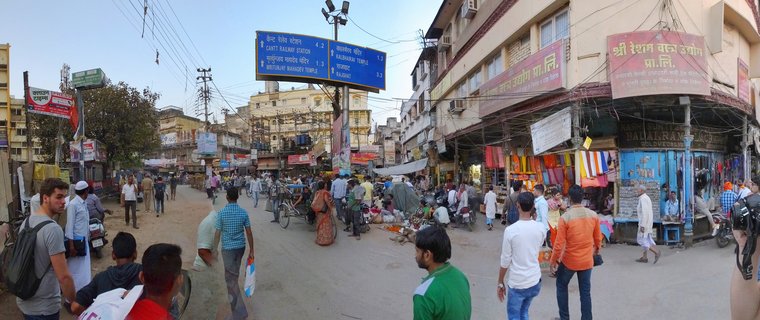Wide street scene in Varanasi showing pedestrians, cycle rickshaws, multi-story buildings with textile shops, and a blue directional sign in Hindi and English pointing to temples and railway station