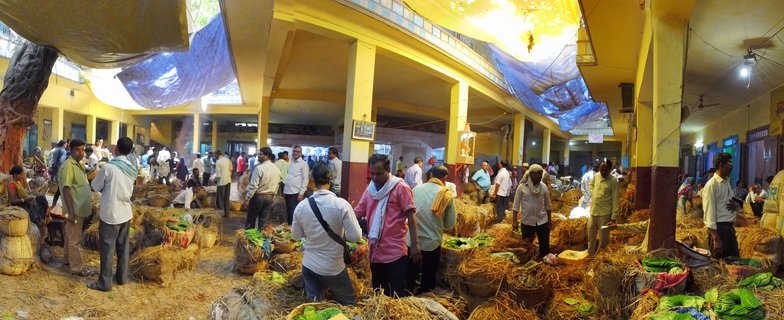 Busy indoor wholesale market in Varanasi, India showing vendors and buyers among woven baskets filled with green betel leaves, yellow pillars, and blue tarps overhead